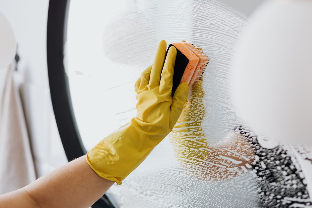 Personne anonyme portant des gants en caoutchouc jaunes et utilisant une éponge simple pour nettoyer le miroir dans la salle de bain